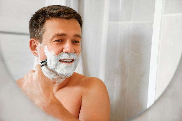 A man shaves his face with a safety razor and looks in a round mirror