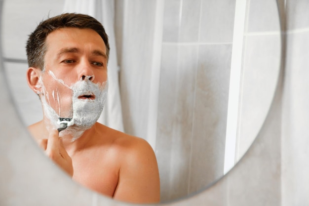 Photo a man shaves his face with a safety razor and looks in a round mirror