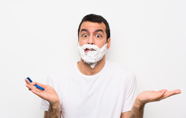 Man shaving his beard over isolated white wall having doubts while raising hands