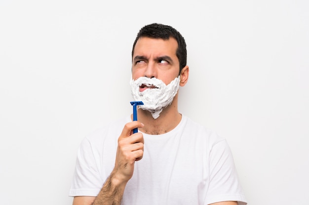 Man shaving his beard over isolated white wall with confuse face expression