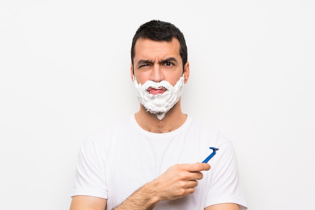 Man shaving his beard over isolated white wall with sad and depressed expression