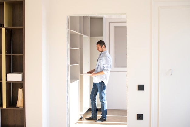 A man in a shirt stands in the apartment and collects shelves in the closet