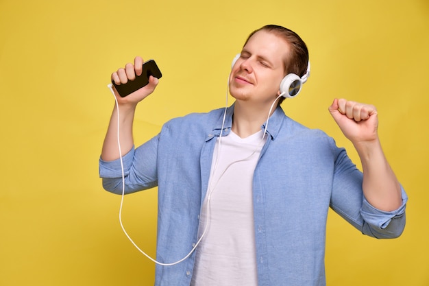 man in shirt on yellow, closing his eyes, listens to music in white headphones from smartphone.