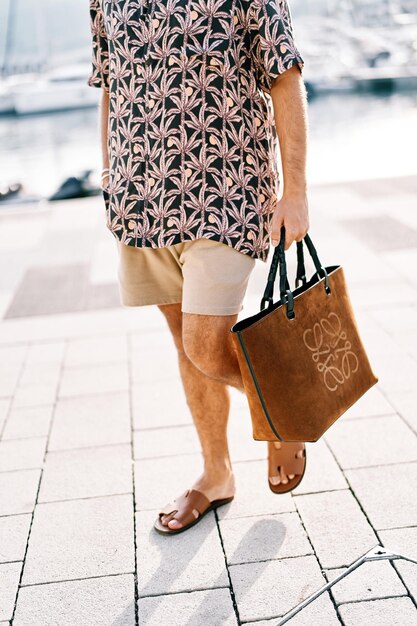 Foto uomo in pantaloncini con una borsa da spiaggia cammina sulle piastrelle nel cortile tagliato senza volto