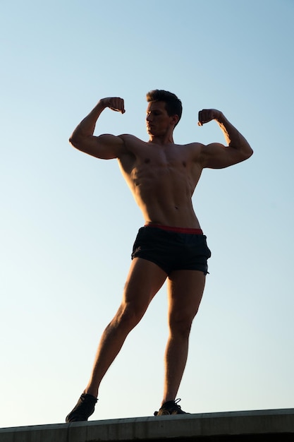 Man showing fit body on blue sky. Bodybuilder posing with muscular hands, biceps and triceps. Athlete standing high on concrete surface. Success and future concept. Fitness and sport.