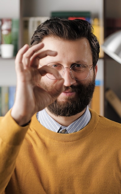 Man shows OK. Positive smiling handsome bearded man in glasses in office looks at camera and shows OK gesture with his fingers. Close-up view