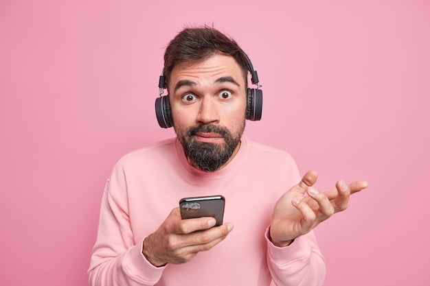  man shrugs shoulders doesnt know how download new application on phone uses digital devices in modern life wears wireless headphones on ears poses against pink wall