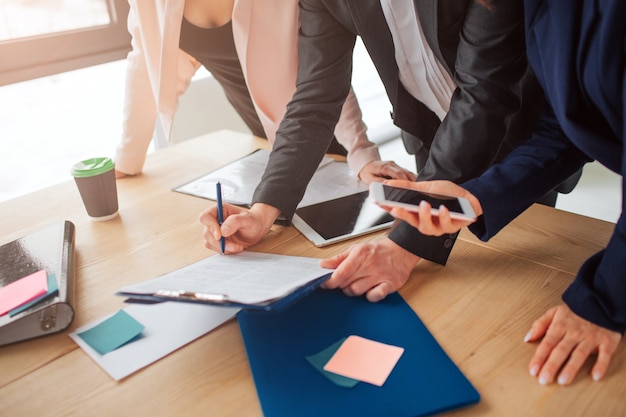 Man signing papers on plastic tablet. Woman's hand hold phone. Another woman lean to table. They work together in one team.