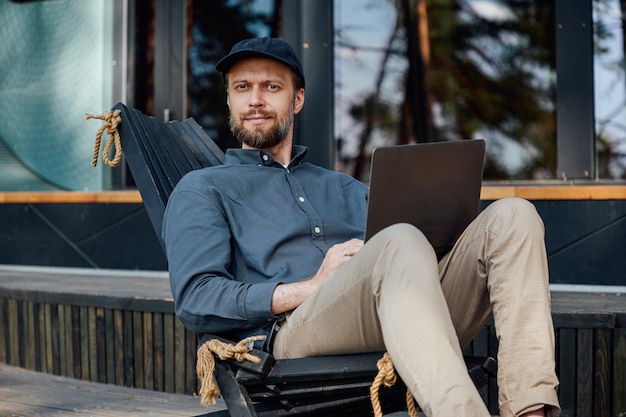 a man sits against the backdrop of panoramic windows of a country house works in a laptop