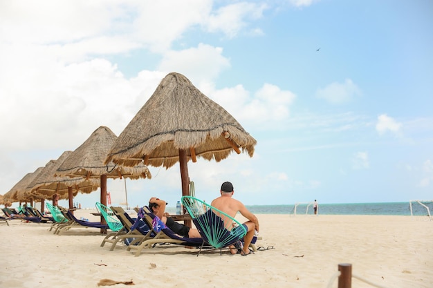 A man sits on a beach with a straw umbrella.