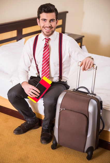 A man sits on a bed and holds his luggage.