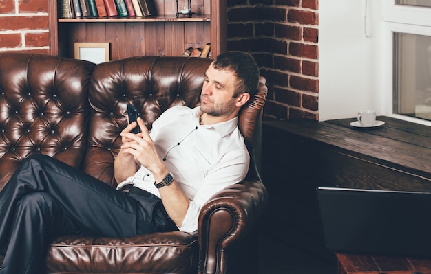 A man sits on a comfortable leather sofa and holds the phone in his hands. Businessman is resting from work behind laptop