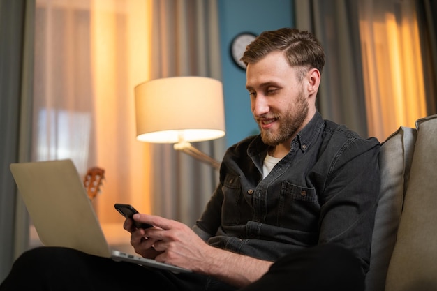 A man sits on couch in the living room in the evening in front of a laptop screen standing on his la