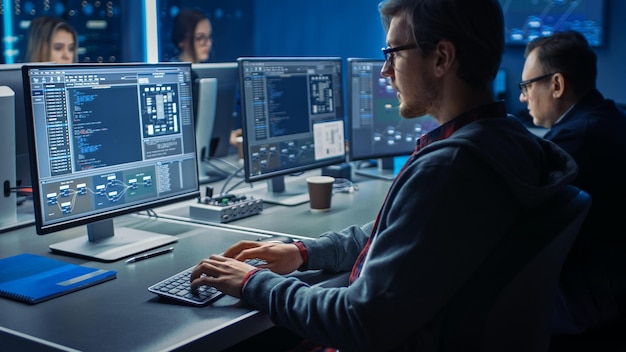 a man sits at a desk in front of two computer monitors, one of which says code on the screen.