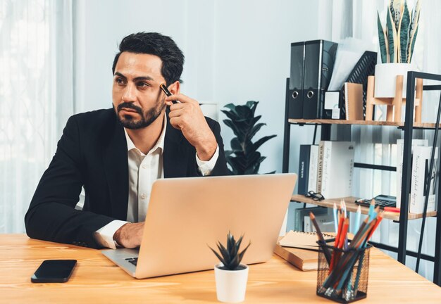 A man sits at a desk with a laptop and a phone in his hand.