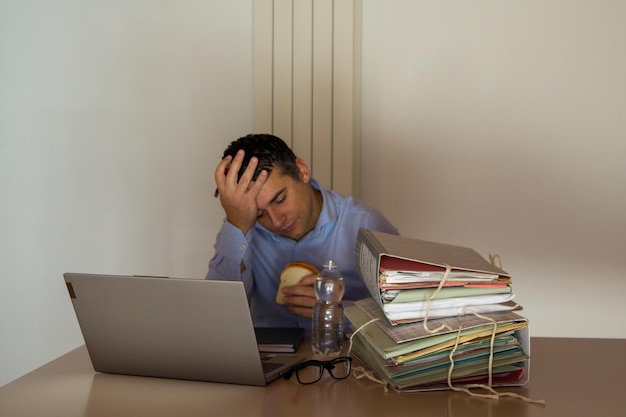 A man sits at a desk with a stack of folders and a laptop.