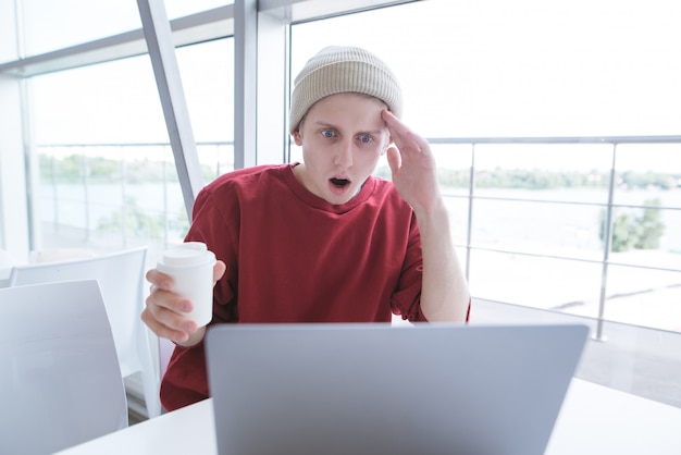 man sits in light cafe with a glass of coffee in hands and looks at laptop screen