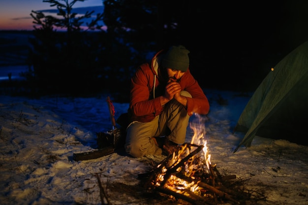 A man sits near a campfire warming himself by the fire in a winter forest at sunset