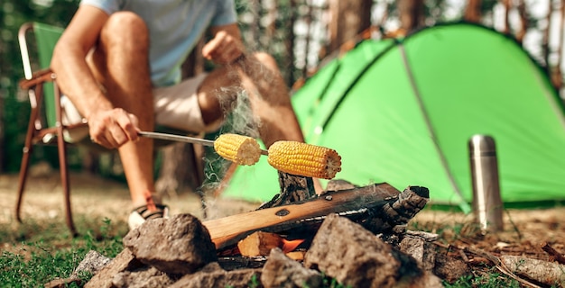 A man sits near a tent by a campfire and fries corn on a skewer\
in a pine forest for the weekend. camping, recreation, hiking.