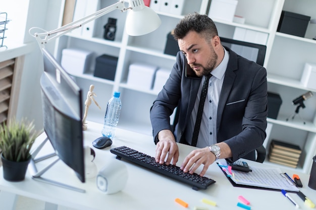 A man sits in the office at the desk, supports the phone with his shoulder and prints on the keyboard.