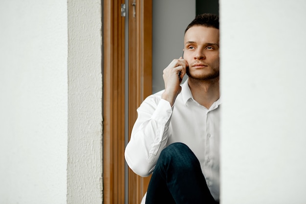 A man sits on an open window looks at the street and talks on the phone