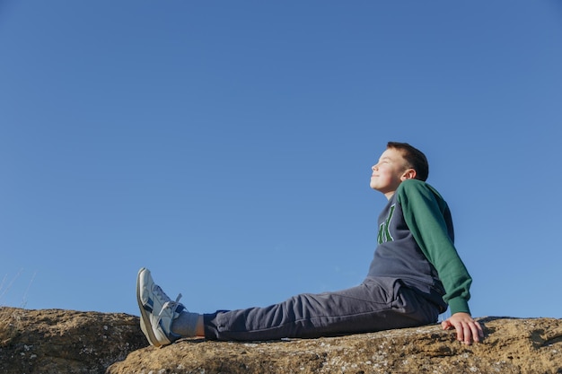 A man sits on a rock in the sun and looks up at the sky.