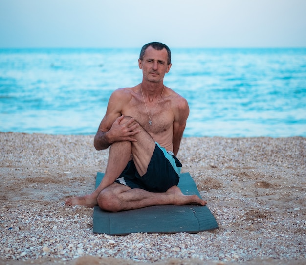 A man sits on a rug, stretches and holds his leg with his hand and looks forward