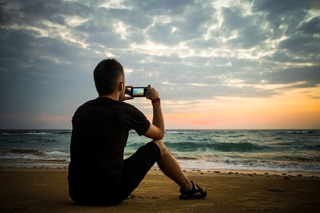 男は砂の上に座って、太陽が地平線から海に昇るときにビデオを撮ります。携帯電話で夕日の写真を撮る男。孤独、孤独、荒廃、孤独