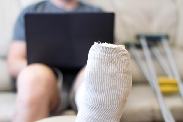 A man sits on a sofa in a cast with a laptop a leg in a cast\
lies on a chair