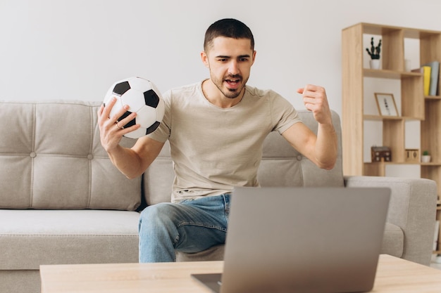 A man sits on a sofa in the living room holding a ball and cheers for the football team watching a match on a laptop