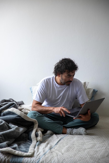 man sitting on a bed typing on a computer, man in his bedroom typing an email on his laptop