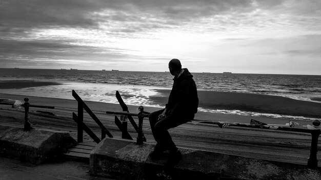 Foto uomo seduto su una panchina mentre guarda il mare contro il cielo