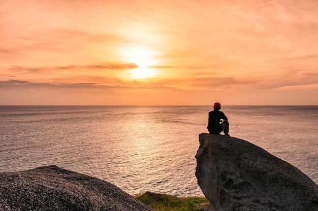 Человек сидит на большой скале с осмотром достопримечательностей на закате на море