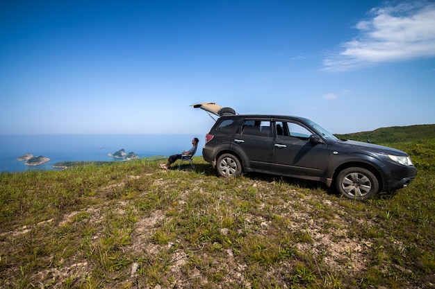 Man sitting at black car's shadow at the top of the hill at sea coast