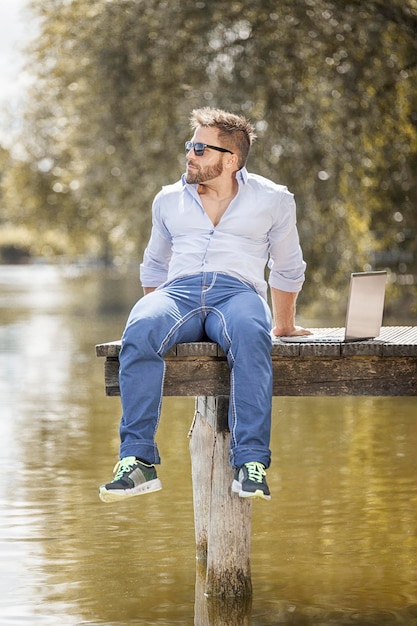 man sitting by laptop on jetty over lake