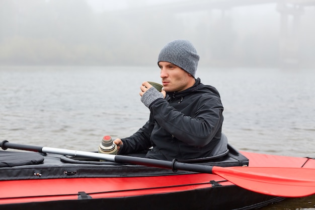 Man sitting in canoe, resting after active paddling on kayak,\
drinking coffee from thermos, looking at beautiful nature, wearing\
black jacket and cap, sitting in middle of lake