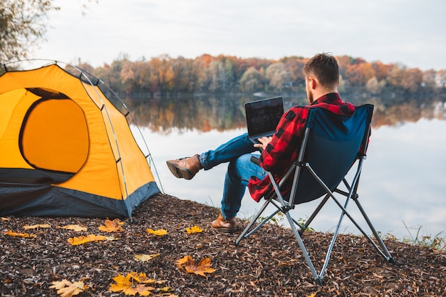 Man sitting in chair near autumn lake working on laptop freelance camping concept