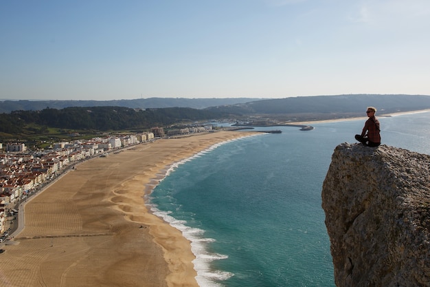 海のビーチと小さな町を上から見ている崖の上に座っている男