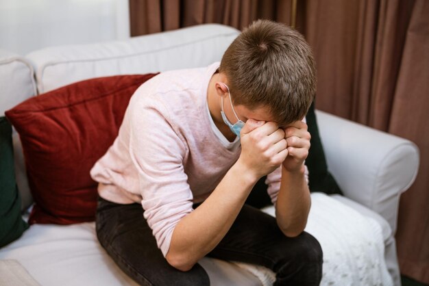 Photo a man sitting on the couch holds his head with his hands in a protective mask