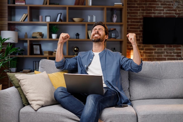 Man sitting on couch working on laptop receiving bank loan approval notification email feeling successful clenching fists making yes gesture Sincere emotions victory result achievement concept