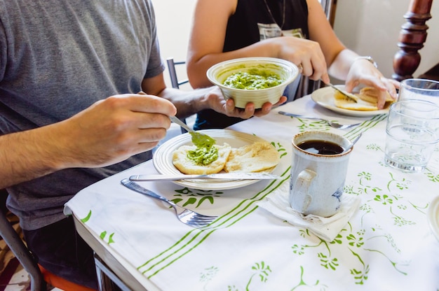 man sitting at the dining table next to his girlfriend eating and filling an arepa with guacamole