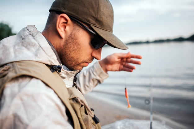 Photo man sitting at the edge of water and looking down. he holds spoon with bait. guy is preparing to fishing.