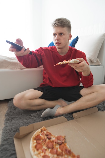 man sitting on the floor in a room with a box of pizza and switching tv channels