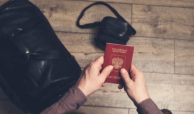 Man sitting holds Russian travel passport in hands with leather bag and photo camera on the floor Travel and tourism concept