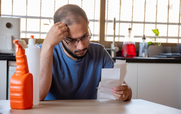 Photo man sitting in the kitchen holding receipt feeling frustrated due inflation and increase of daily needs supplies for house with detergents in front of him
