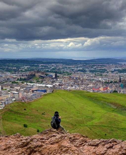 Фото Человек сидит на горе на фоне городского пейзажа и облачного неба