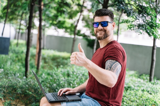 Man sitting in a park working remotely with his laptop and gesturing with his finger raised in signal that everything is okay