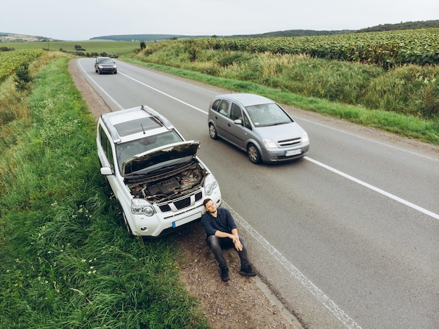 壊れた車の近くの道路に座って助けを求めて車を止めようとしている男