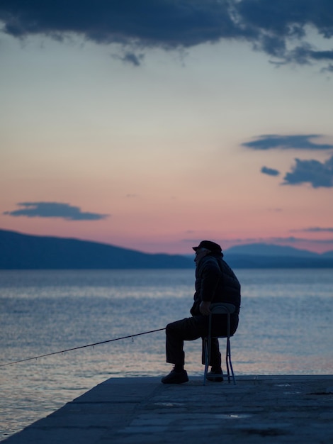 Photo man sitting on shore against sky during sunset