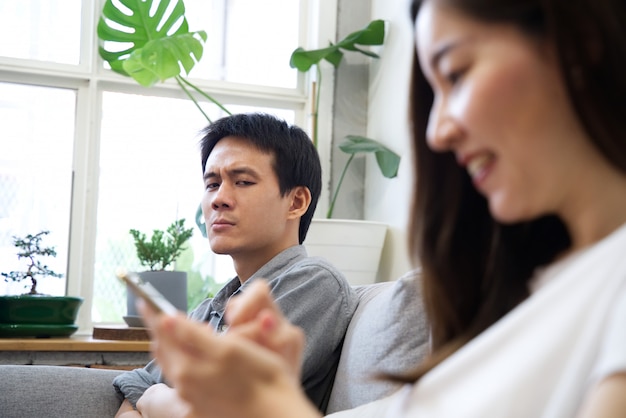 A Man sitting on sofa is feeling unhappy with his girlfriend while she enjoys using phone